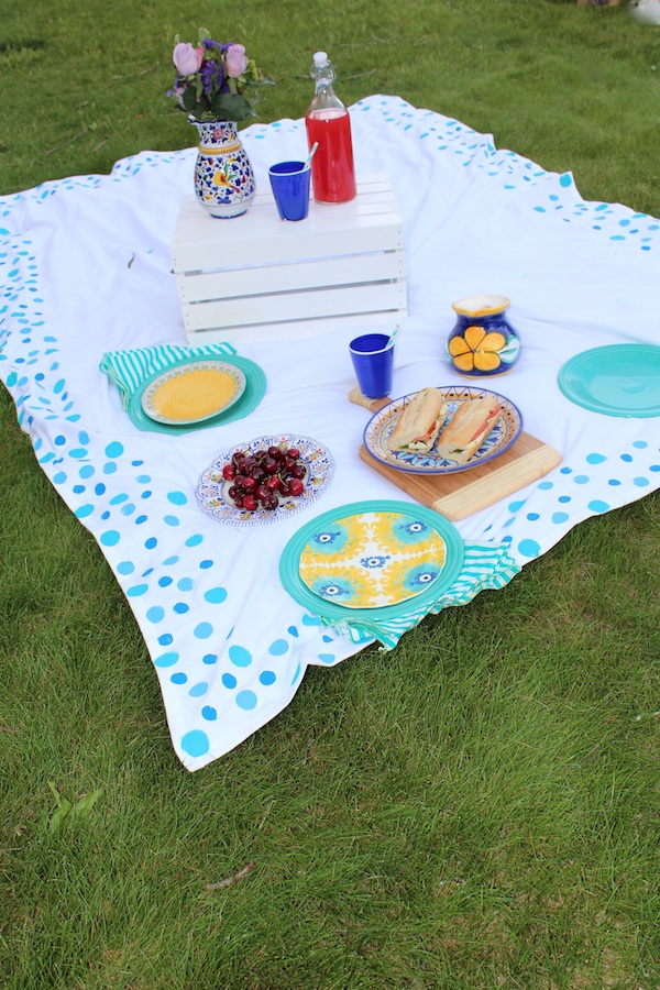 White picnic blanket with blue spots all around, set up for a picnic outside on the grass.