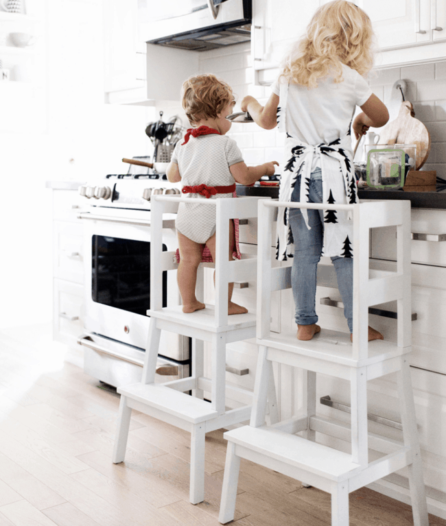 2 children on white high learning towers helping in the kitchen.