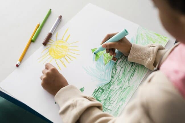 Child drawing a sun and grass on a white sheet of paper with crayons.
