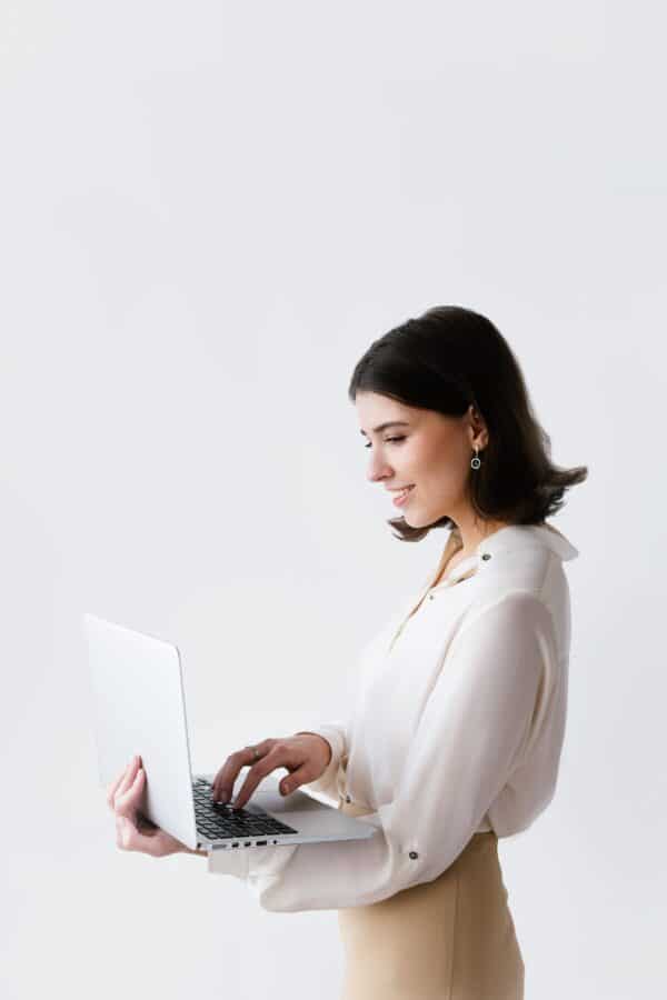 Woman standing holding a laptop wearing a cream blouse and tan colored skirt.